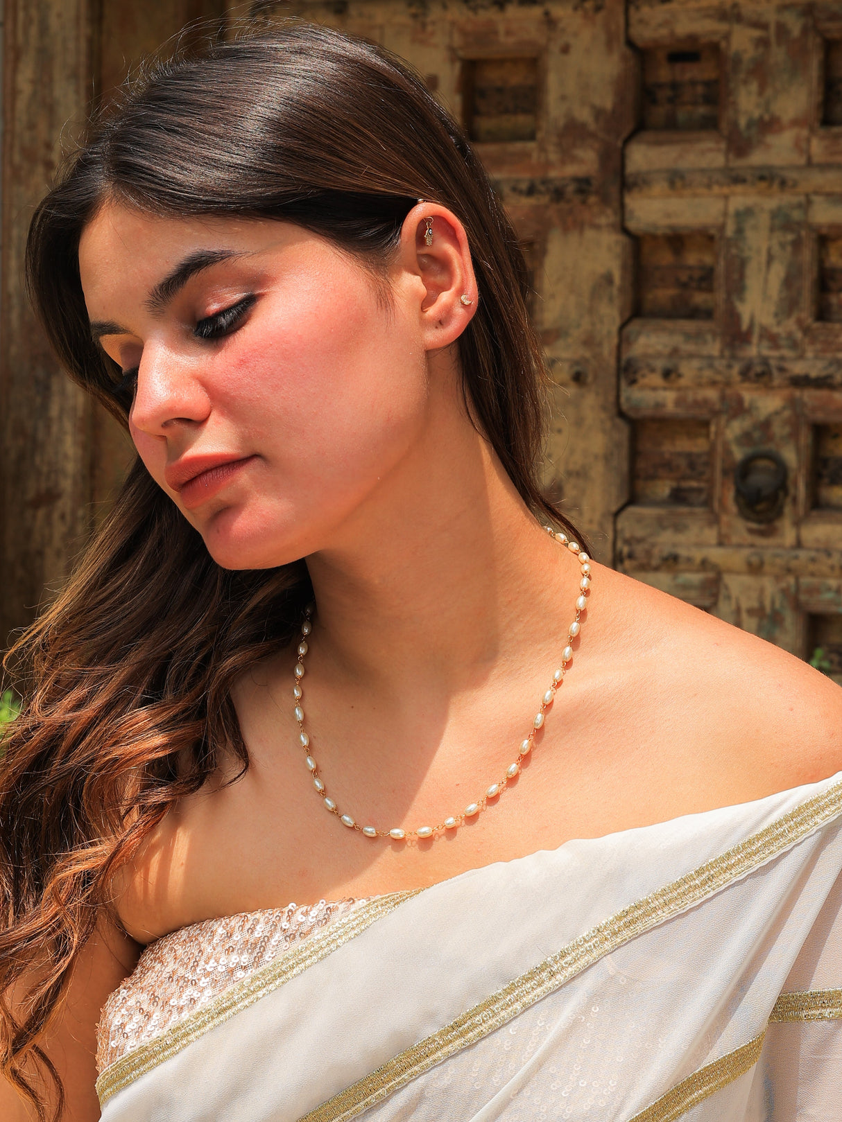 Woman wearing a white saree with gold border and pearl necklace against a textured wall.
