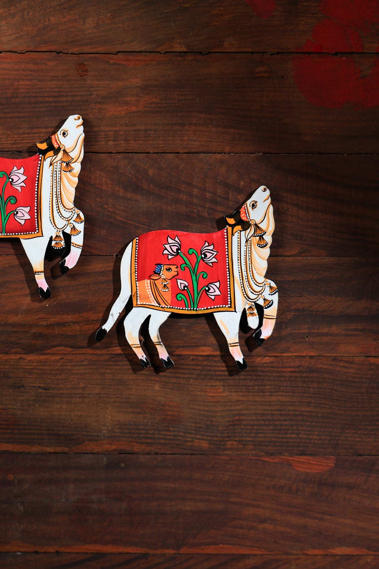 Two hand-painted MDF cutouts of cows with floral patterns on a red background, placed on a wooden surface.