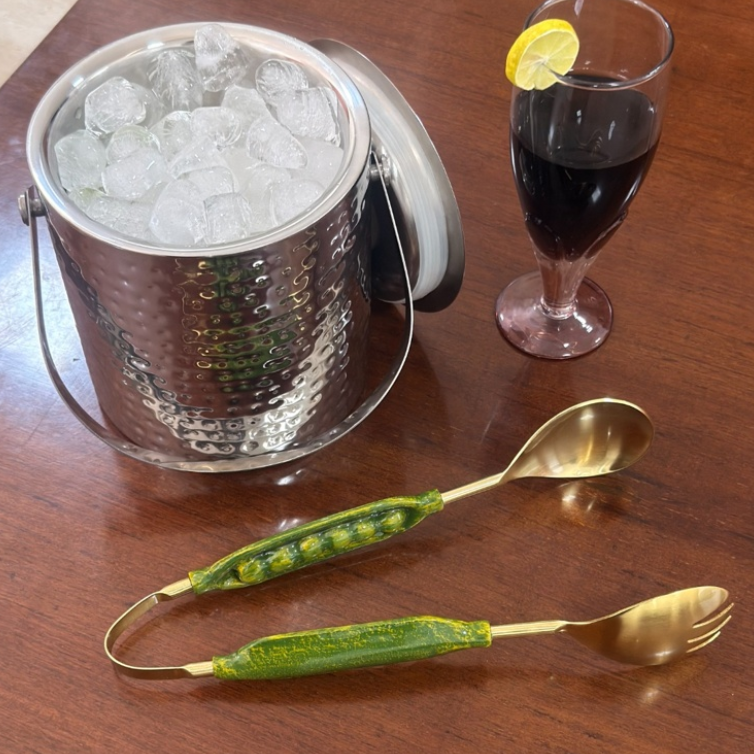 Gold tongs with green handles, ice bucket, and glass of dark liquid on a wooden surface